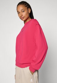 Sweatshirt fúcia com mangas longas, gola canelada e detalhe do logótipo no peito. Usado com calças de corte descontraído em tom claro.