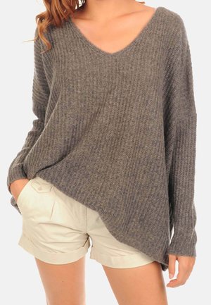 Femme portant un pull ample gris à côtes avec un col en V et des manches longues, ainsi qu'un short beige à revers, une main dans la poche.