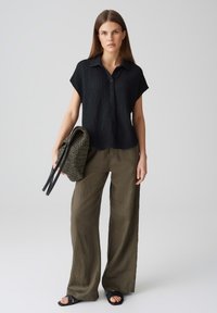 Camicia nera in tessuto testurizzato a maniche corte con colletto, abbinata a pantaloni larghi in lino verde oliva e una borsa intrecciata verde oliva con manici neri.
