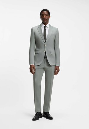 Mężczyzna ubrany w jasnoszary garnitur z dwoma guzikami, białą koszulę, czarny krawat oraz czarne eleganckie buty stoi na gładkim białym tle.