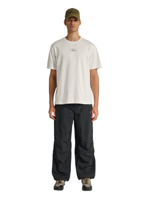 Mand iført hvid HALO T-shirt, sorte løse bukser, beige og sorte vandresko og olivengrøn HALO kasket, stående med fronten mod frem.