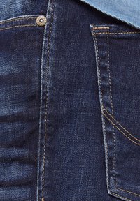 Primo piano di jeans in denim blu scuro che mostrano le cuciture, la tasca posteriore e il dettaglio del bottone in metallo con marchio.