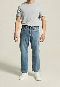 Homme portant un t-shirt gris clair à manches courtes, un jean droit bleu et des baskets blanches, debout devant un fond beige uni.