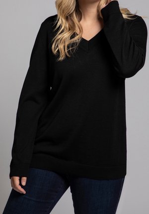 Femme portant un pull noir à col en V manches longues et un jean bleu foncé, avec des cheveux blonds détachés, debout devant un fond gris.