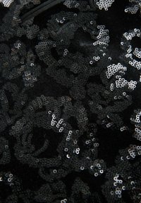 Zwarte stof versierd met intricated zilveren pailletten, met een bloemenpatroon. De textuur is soepel met een glanzende reflecterende kwaliteit.