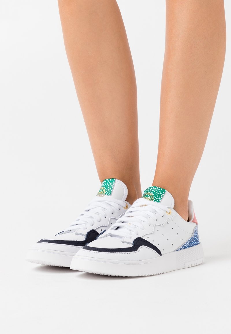 Vita idrottsskor med marinblå accent, grön prickig tunga och en texturerad, flerfärgad häl. Snörningsdesign med perforeringar.