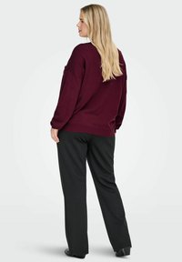 Burgundowa, oversize sweter z opadającymi ramionami, prążkowanym wykończeniem i długimi rękawami, w połączeniu z czarnymi spodniami o szerokich nogawkach i płaskimi butami.