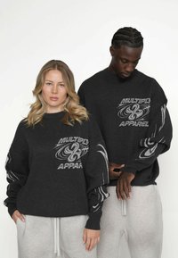 Hanorace de culoare gri închis cu manșete ribbed și un grafic mare cu textul "MULTIPLY APPAREL." Modelul poartă pantaloni de trening gri cu șnur.