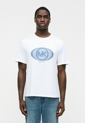 Mann trägt ein weißes Michael Kors T-Shirt mit blauem MK-Logo und blaue Jeans, steht vor einem schlichten weißen Hintergrund.