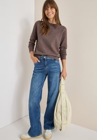 Brązowy sweter, szerokie niebieskie dżinsy, kremowa torba na ramię i jasne sneakersy. Dżinsy mają wyblakłe wykończenie i zapięcie na guzik.