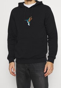 Schwarzer Hoodie mit einem bunten bestickten Kolibri-Design auf der Brust, ausgestattet mit einer Kängurutasche sowie gerippten Bündchen und Saum.