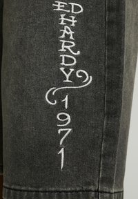 Tissu en denim noir avec texte brodé blanc « ED HARDY 1971 » disposé verticalement avec un motif tourbillonnant décoratif.