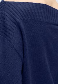 Gros plan d’un pull bleu marine à épaules dénudées montrant un col côtelé et un tissu en maille texturé sur l’épaule et le haut de la poitrine.