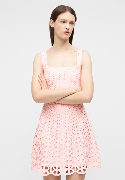 Νεαρή γυναίκα στέκεται με σταυρωμένα χέρια φορώντας ροζ φόρεμα χωρίς μανίκια από δαντέλα με τετράγωνο ντεκολτέ και φούστα με σχέδια, μπροστά σε απλό φόντο.