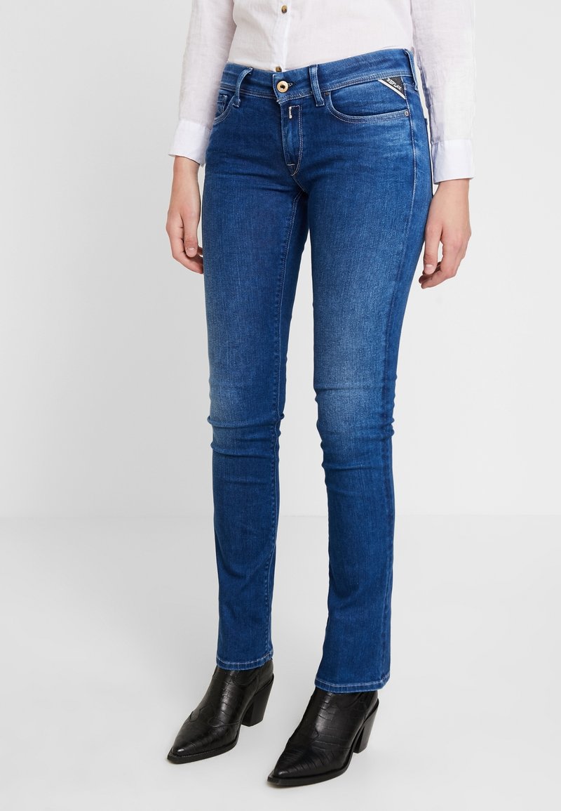 Femme portant un jean bleu slim et des bottines noires à talons, debout devant un fond blanc uni.