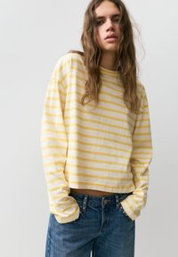 Femme aux longs cheveux ondulés portant un t-shirt à manches longues rayé jaune et blanc et un jean bleu, debout les mains dans les poches.