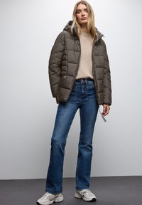 Padded oliwkowa kurtka z kapturem, beżowy sweter i dopasowane niebieskie dżinsy. Szare sneakersy z akcentami dopełniają całość.