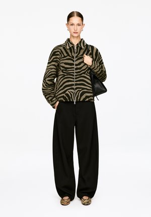 Kurtka polarowa w wzór zebry w kolorach oliwkowym i czarnym z zamkiem błyskawicznym, zestawiona z czarnymi spodniami o szerokich nogawkach oraz wsuwanymi butami w paski.