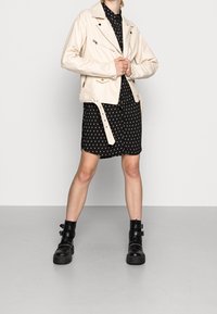 Krémová faux kožená biker bunda se zipovými detaily, nošená přes černé šaty s puntíky. Černé masivní kotníkové boty na přezky doplňují celý outfit.