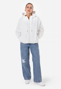 Felpa bianca con zip e cappuccio ampio, abbinata a jeans a vita alta in blu chiaro con un motivo strappato e scarpe da ginnastica bianche.