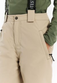Beige waterdichte broek met een gladde textuur, voorzien van zijzakken, een verstelbare riem en een knoopsluiting. Gecombineerd met een groene top.