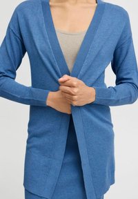 Blauwe cardigan van zachte brei materiaal, met een V-hals, lange mouwen en een open voorzijde met ribbels aan de randen.