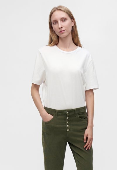 Jeune femme aux longs cheveux blonds portant un t-shirt blanc uni et un pantalon taille haute vert olive avec des boutons, debout avec une main dans la poche.