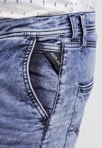Jean en denim de lavage bleu moyen avec une texture délavée, doté d'une poche latérale avec une étiquette noire sur laquelle est inscrit "REPLAY" et des coutures détaillées.