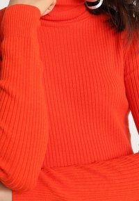 Pull à col roulé côtelé de couleur orange vif, avec des manches longues et une coupe ajustée. Le matériau semble doux avec une texture visible.