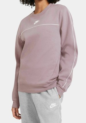 Person iført mauve Nike sweatshirt med hvide piping og lysegrå Nike træningsbukser med hænder afslappet ned langs siderne.