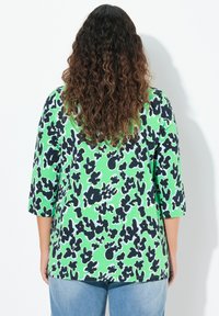 Zielona bluzka z czarno-białym abstrakcyjnym wzorem, z rękawami do 3/4, okrągłym dekoltem i luźnym krojem, w połączeniu z niebieskimi dżinsami.