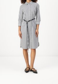 Pruhované šaty košilového stylu s límečkem, třičtvrtečními rukávy, staženým pasem s páskem s logem a rozšířeným lemem. Nosené s černými balerínami.