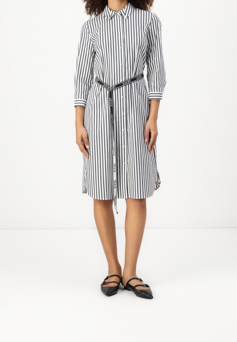 Pruhované šaty košilového stylu s límečkem, třičtvrtečními rukávy, staženým pasem s páskem s logem a rozšířeným lemem. Nosené s černými balerínami.