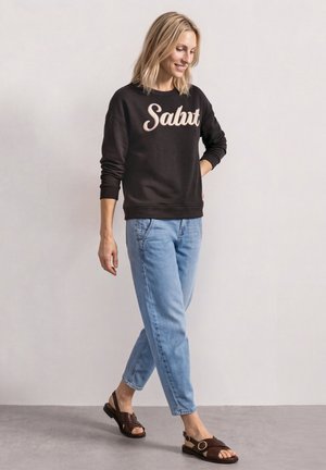 Lyshåret kvinde, der går, iført en sort sweatshirt med teksten "Salut", lyseblå jeans og mørkebrune åbne sandaler mod en ensfarvet lys baggrund.