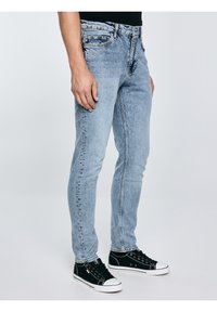 Mees, kes kannab helesiniseid kitsaid teksapükse ja musti madala säärega tossusid, seisab ühevärvilise hele tausta ees.