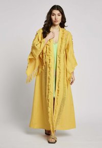 Lange gele kimono met franjes. Lichtgewicht stof, open voorkant en brede mouwen. Gemaakt over een groene kledingstuk en gecombineerd met sandalen.
