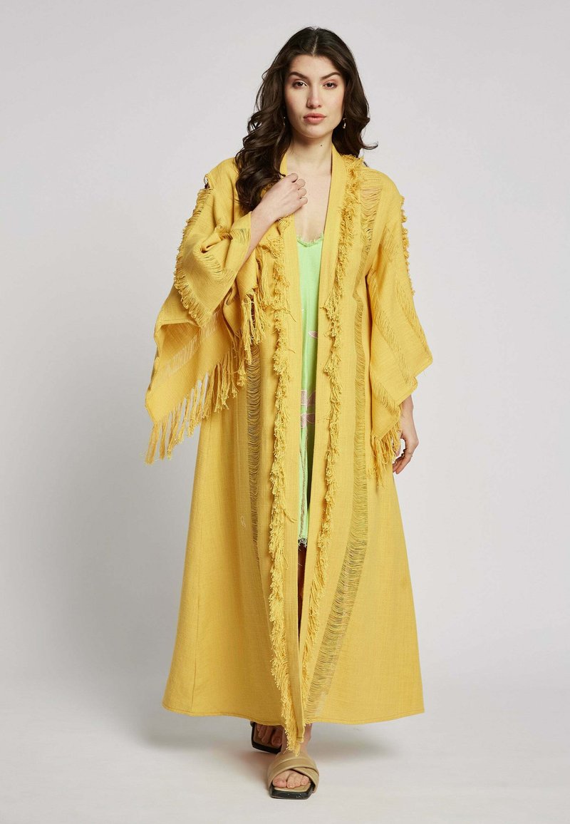 Lange gele kimono met franjes. Lichtgewicht stof, open voorkant en brede mouwen. Gemaakt over een groene kledingstuk en gecombineerd met sandalen.