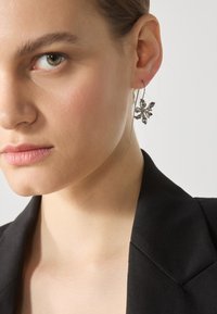 Cercei argintii în formă de floare cu pietre semiprețioase transparente, având un design cu cârlig lung. Purtați la urechea stângă, asortați cu un sacou închis la culoare.