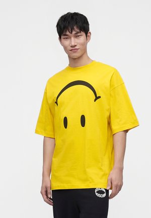 Jeune homme aux cheveux courts noirs portant un t-shirt oversize jaune vif avec un grand motif visage souriant noir.