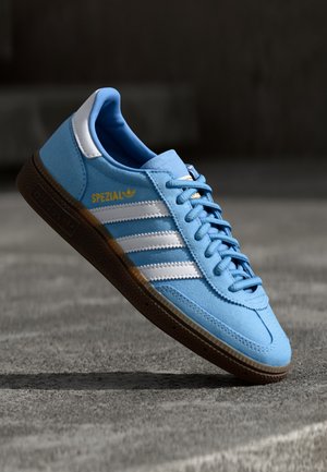 Lyseblå Adidas Spezial sneaker med hvide striber, brun sål og guldskrift på en grå overflade i blødt lys.