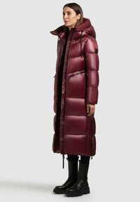 Bordeaux pufferjack met een capuchon, lange stijl, glanzende textuur, ritssluiting aan de voorkant en twee zijzakken. Draagt met zwarte haklaarzen.