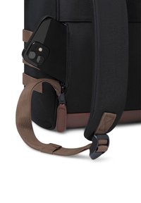 Sac à dos en tissu noir avec une sangle marron, doté d'une poche zippée contenant un smartphone noir. Accents texturés et quincaillerie robuste.