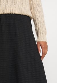 Pletený béžový svetr s texturovanou černou sukní s mřížkovým vzorem. Ruka spočívá na pasové linii sukně.