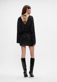 Moteris vilki juodą blizgiu mini suknelę su ilgomis rankovėmis, atvira nugaros dalimi, surišta virvėmis, ir juodas aukštakulnius batus iki kelio.