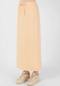 Light peach midi skirt in soft fabric, featuring an elastic waistband with drawstring, side slits, and straight cut hem. Paired with beige sneakers.