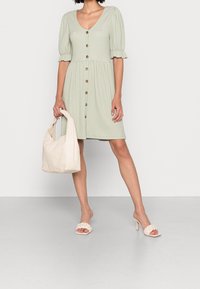 Kobieta w jasnozielonej sukience zapinanej na guziki z bufkami, trzyma torebkę w kolorze kremowym i nosi kremowe sandały na obcasie z plecionki.