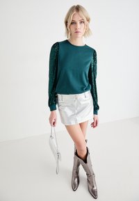 Top de manga larga en color azul verdoso con acentos de lentejuelas, combinado con una mini falda plateada y botas metálicas hasta la rodilla. Se incluye un pequeño bolso plateado.