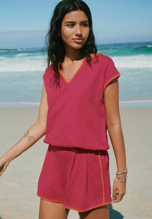 Hosszú, sötét hajú nő rózsaszín ujjatlan felsőt és narancssárga szegélyű rövidnadrágot visel, homokos strandon áll, háttérben hullámokkal.