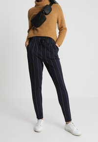 Brown knit turtleneck sweater paired with navy pinstriped trousers and white sneakers. A black crossbody bag sits on the shoulder.