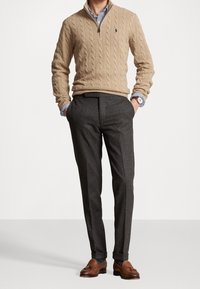 Homme portant un pull beige en maille torsadée, une chemise rayée, un pantalon gris foncé, des mocassins en cuir marron à pampilles, et une montre-bracelet, les mains dans les poches.